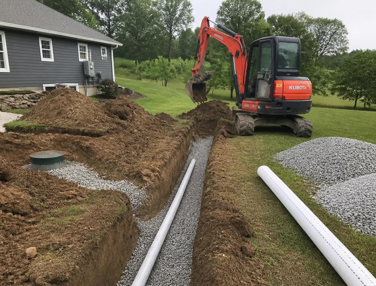 Compact excavator digging a drainfield trench with gravel and perforated pipe beside a home